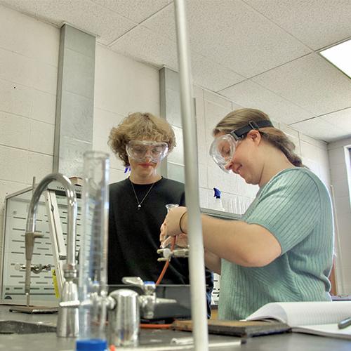 Two individuals wearing safety goggles conducting an experiment together on a lab bench in a science classroom.